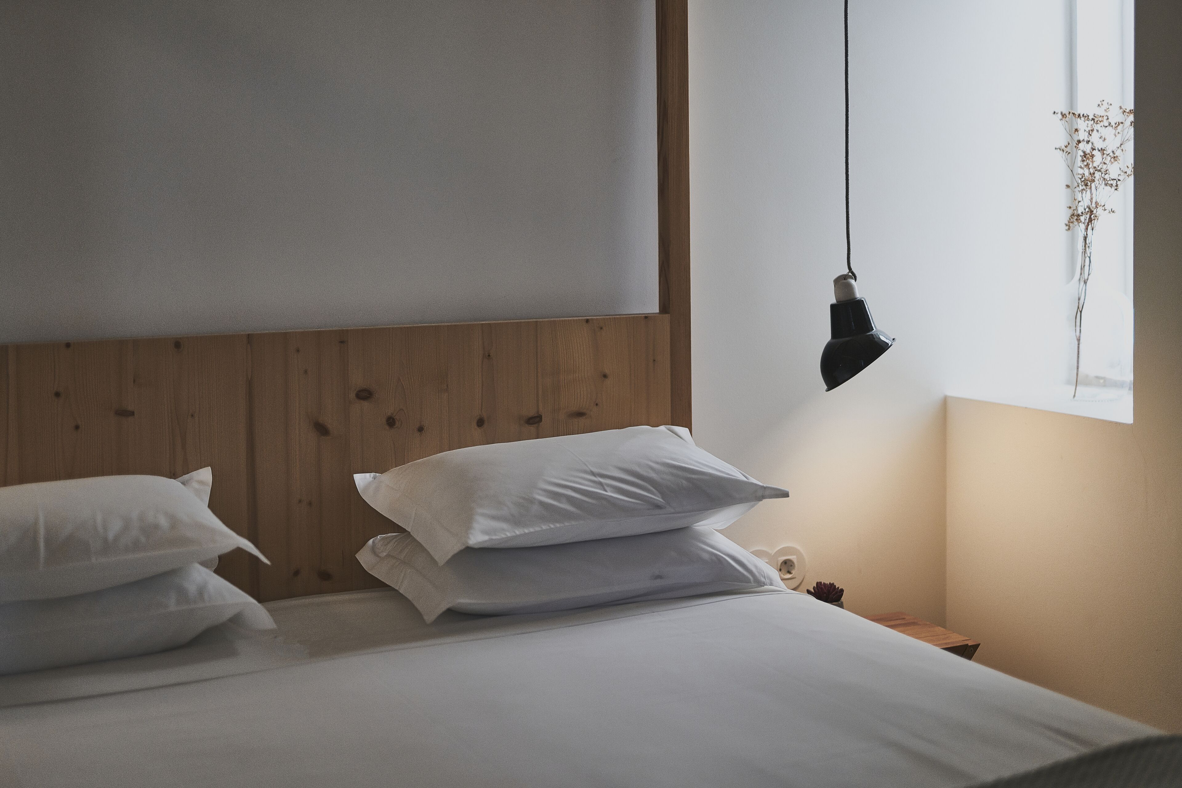 Een netjes opgemaakt bed met witte lakens en kussens, een houten hoofdbord, een hanglamp en een raam met uitzicht.