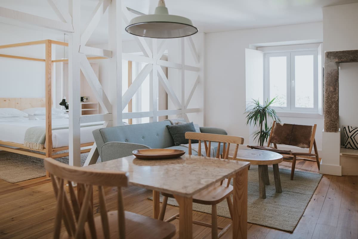 Een moderne, goed verlichte kamer met een houten vloer, een bed met wit beddengoed, een blauwe bank, een eettafel met stoelen en een groot raam dat natuurlijk licht binnenlaat.