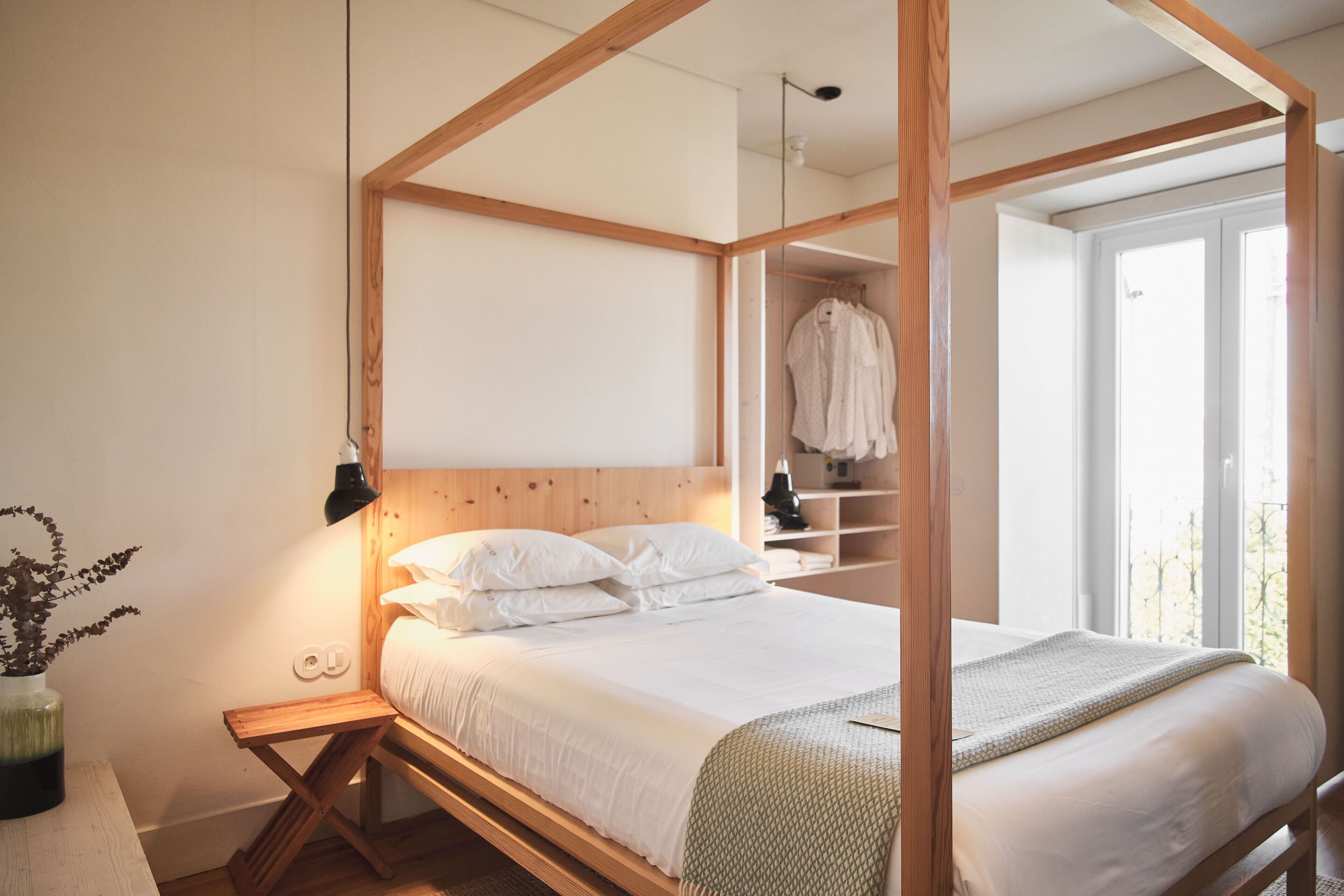 A bedroom with a wooden four-poster bed, a bedside table, and a nightstand. There is a window with a view of greenery outside.