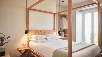 A bedroom with a wooden four-poster bed, a bedside table, and a nightstand. There is a window with a view of greenery outside.