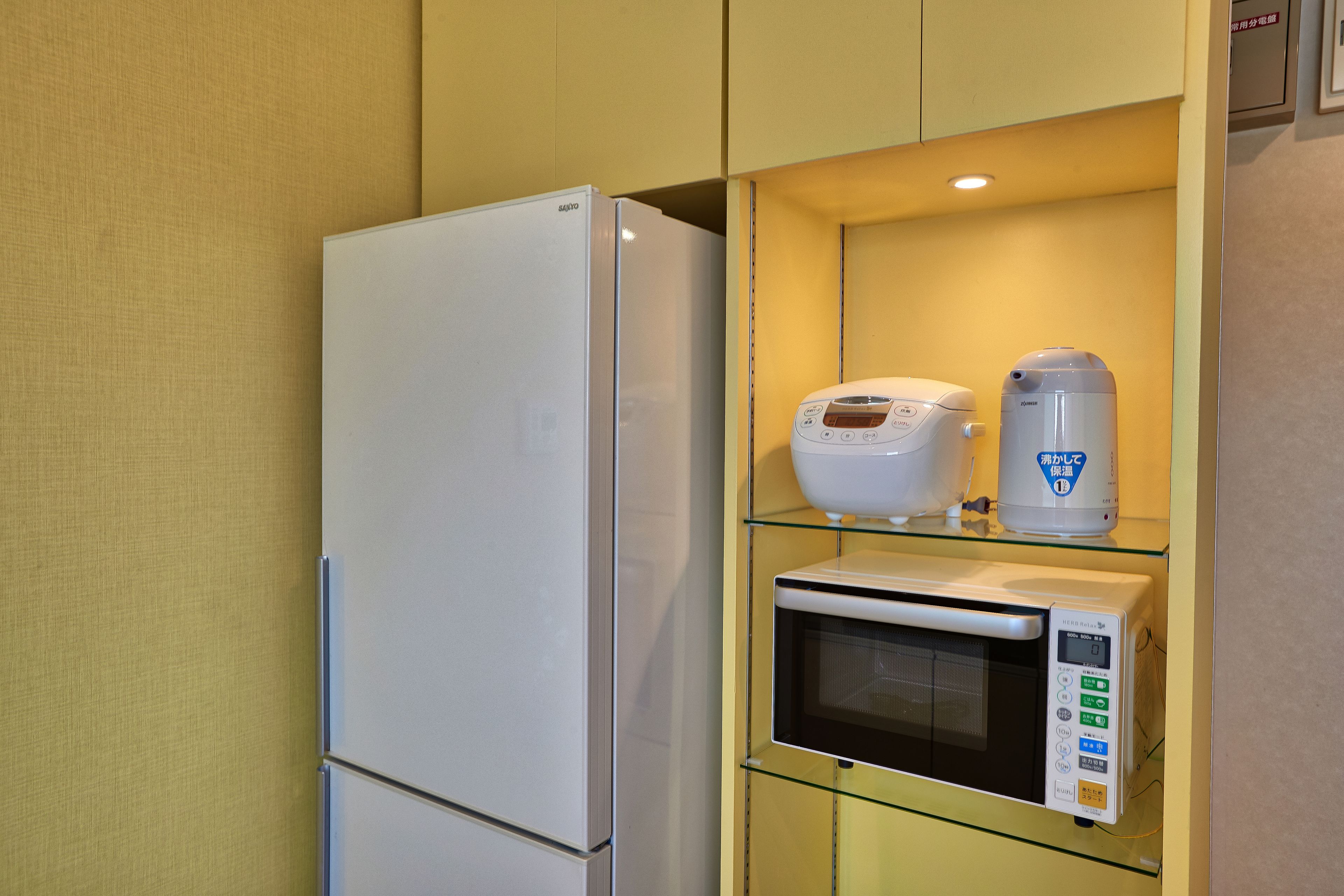 A compact kitchenette with a white refrigerator, a microwave, and a coffee maker on a glass shelf.