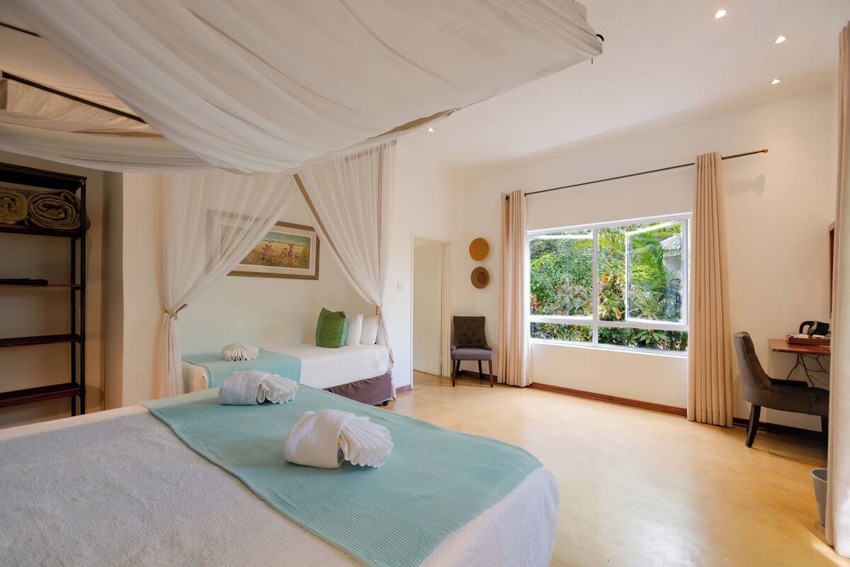A hotel room with a large bed, a desk, a chair, and a window with a view of greenery.