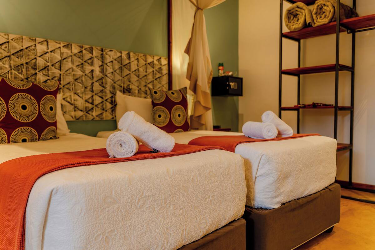 A hotel room with two beds, each with a rolled towel, a red blanket, and patterned pillows.