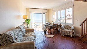 A spacious living area with a sofa, a covered chair, a small table, and a dining table with chairs. There is a large window providing a view of the outside. - LE BELVEDERE AP1059 by RIVIERA HOLIDAY HOMES (Villefranche-sur-Mer)