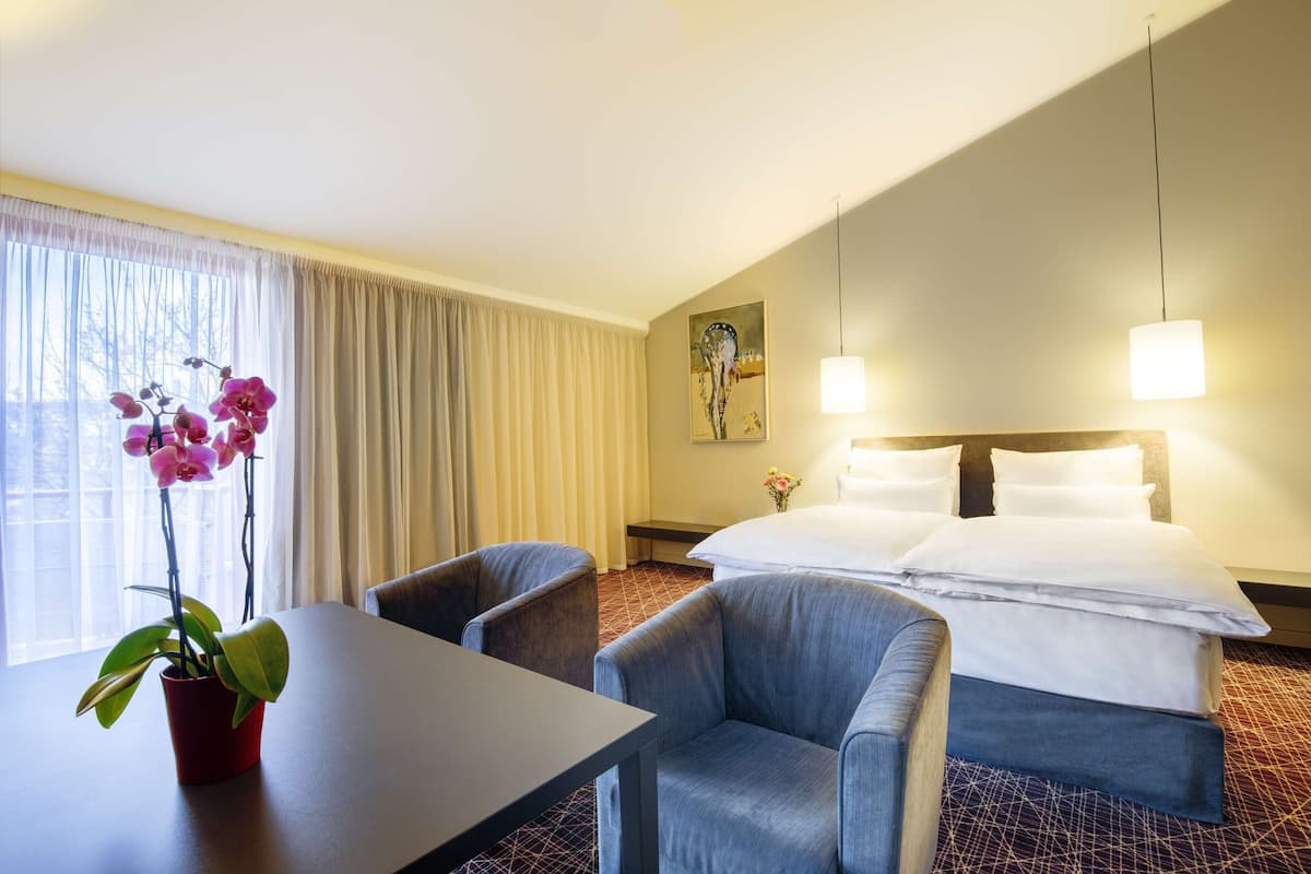 A hotel room with a bed, a desk, two chairs, a potted orchid, and a painting on the wall.