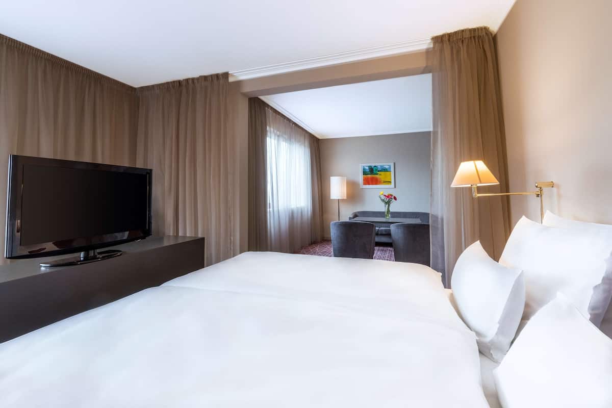 A hotel room with a large bed, a flat-screen TV on a stand, a desk with a chair, and a lamp.
