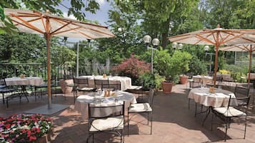 Eine Terrasse im Freien mit runden Tischen und Stühlen, beschattet durch große Sonnenschirme, umgeben von viel Grün und Topfpflanzen.