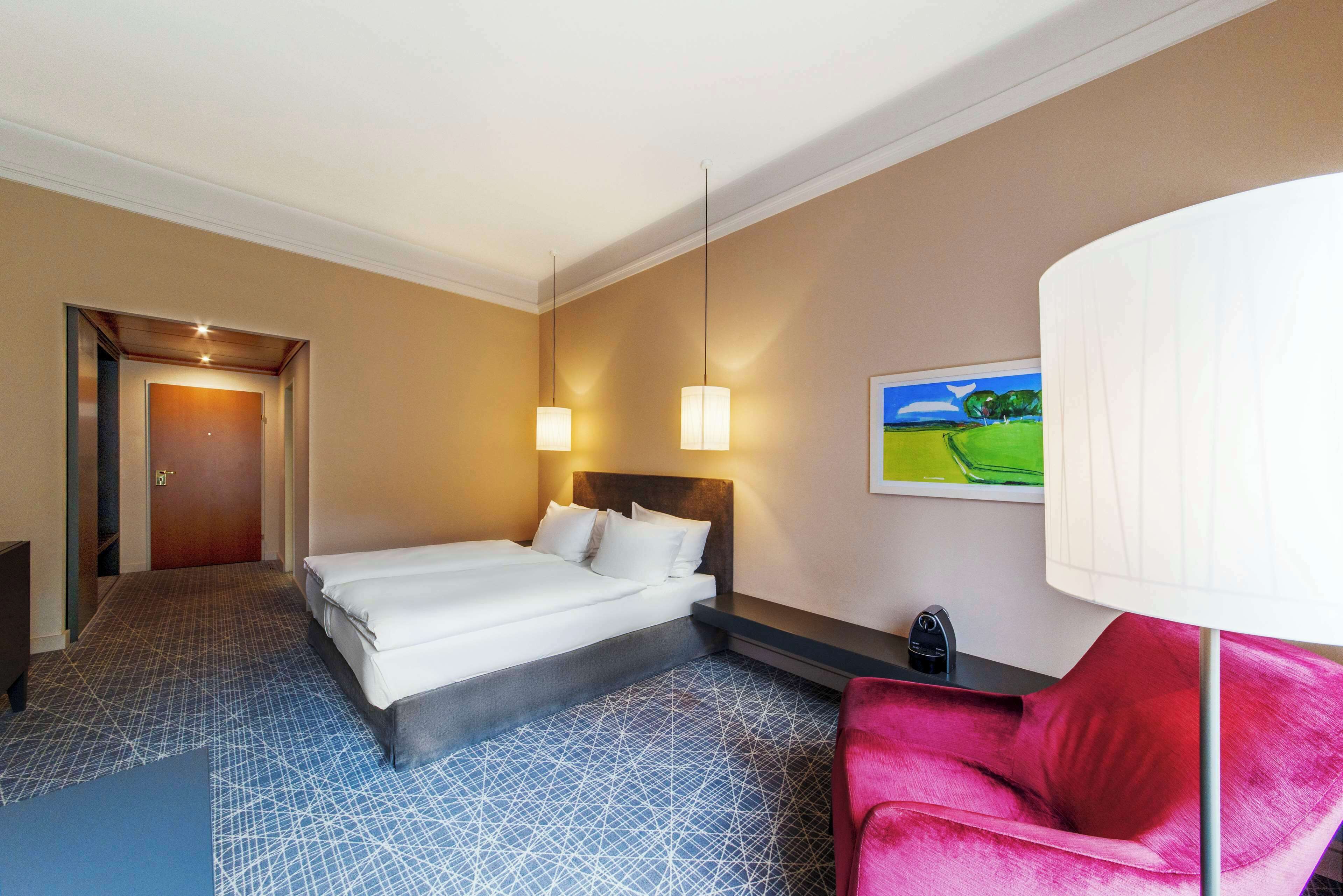 A hotel room with a bed, a desk, a chair, a lamp, and a painting on the wall.
