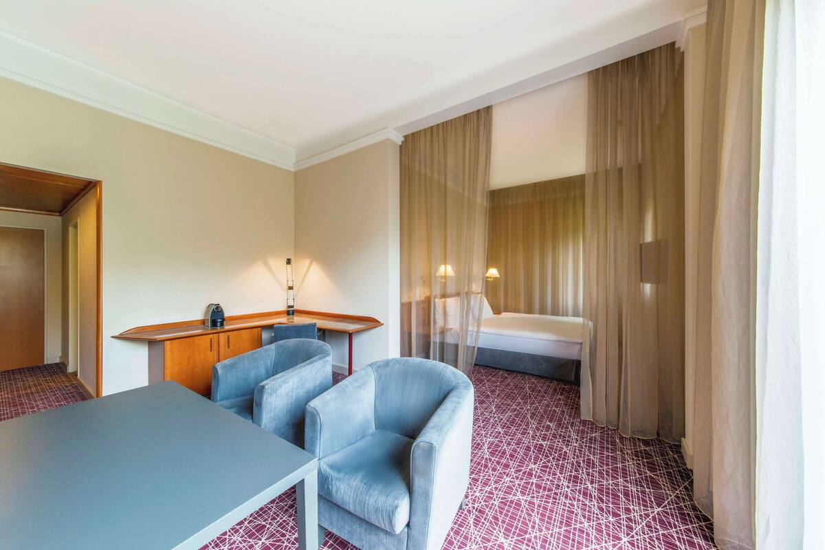 A hotel room with a desk, two chairs, a bed, and a window with curtains.