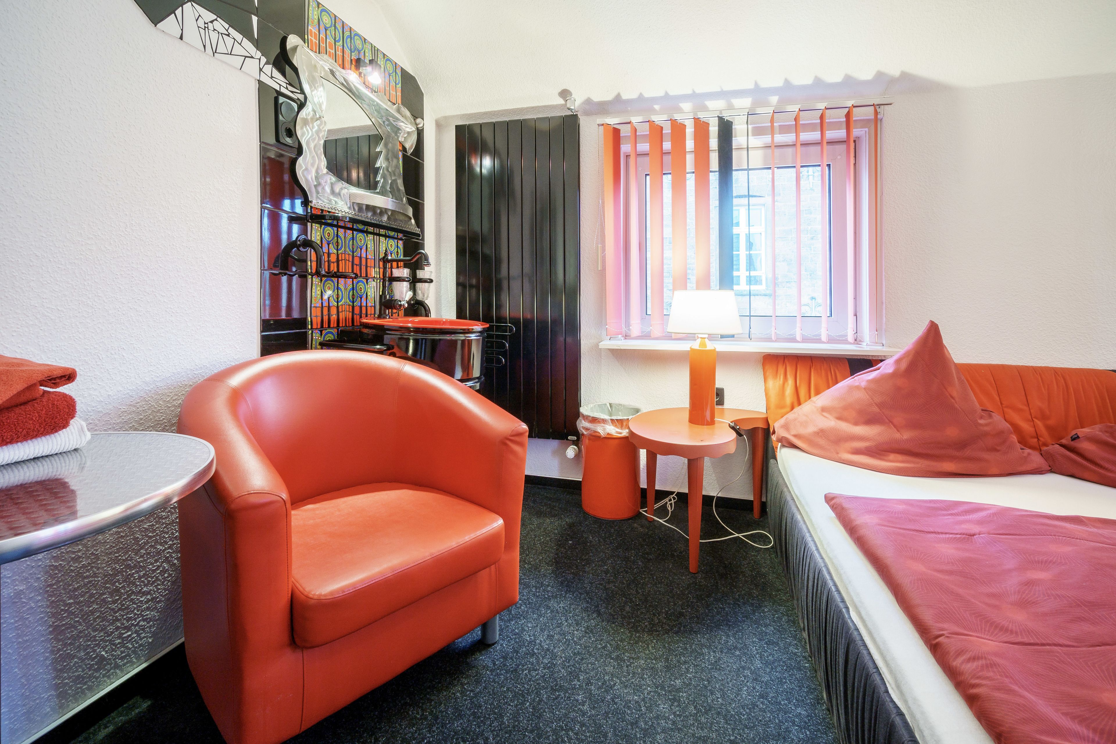 Ein kleines Hotelzimmer mit einem Bett, einem roten Sessel, einem kleinen Tisch, einer Lampe und einem Fenster mit Vorhängen.