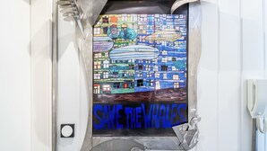 Bany amb mural a la paret que representa un paisatge urbà i un gran cartell que diu 'SAVE THE WHALES'.