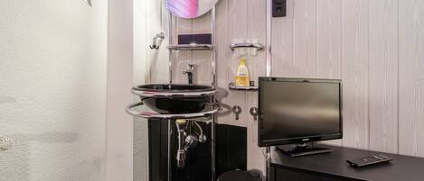 Un baño con lavamanos negro, un espejo, un inodoro, un estante con artículos de aseo y un televisor sobre un pedestal negro.