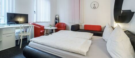 A hotel room with a bed, a desk, a chair, a red sofa, a small table, and a clock on the wall.