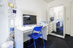 Habitación de hotel pequeña con escritorio blanco, una silla azul, televisor de pantalla plana, espejo y un cubo de basura.