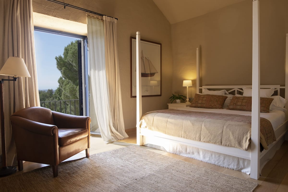 Ein Schlafzimmer mit einem Himmelbett, einem braunen Ledersessel, einem großen Fenster mit Vorhängen und einem gerahmten Bild an der Wand.