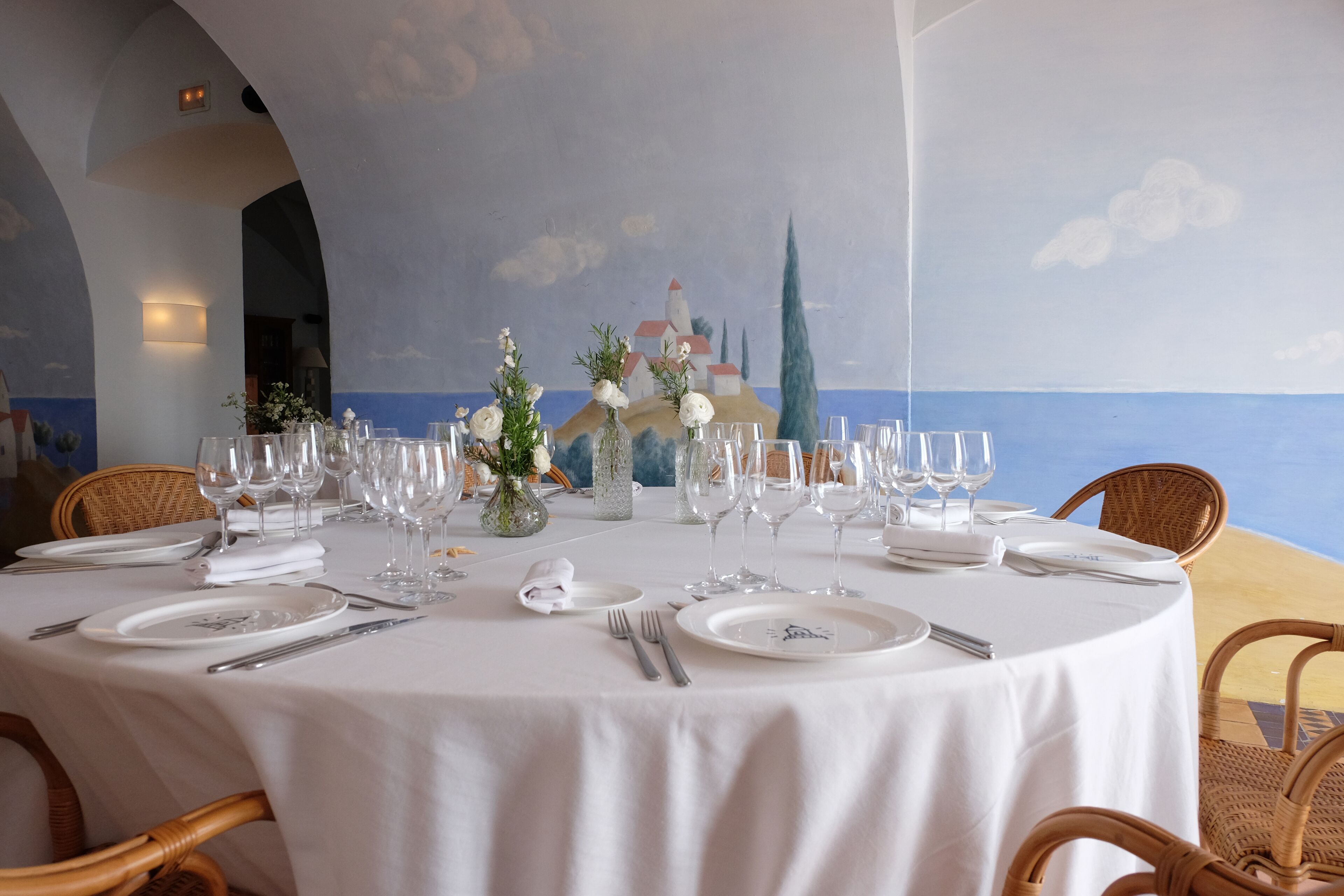 Een ronde eettafel gedekt voor zes personen, met witte tafelkleden, servies en bestek. Voor de tafel hangt een muurschildering van een kustlandschap met gebouwen en een kerktoren.