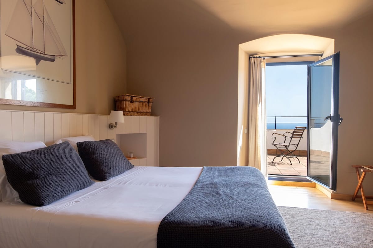 Ein Schlafzimmer mit Bett, Kissen, einem Nachttisch, einer Lampe, einem gerahmten Bild, einem Korbwäsche und einem Balkon mit einem Stuhl.