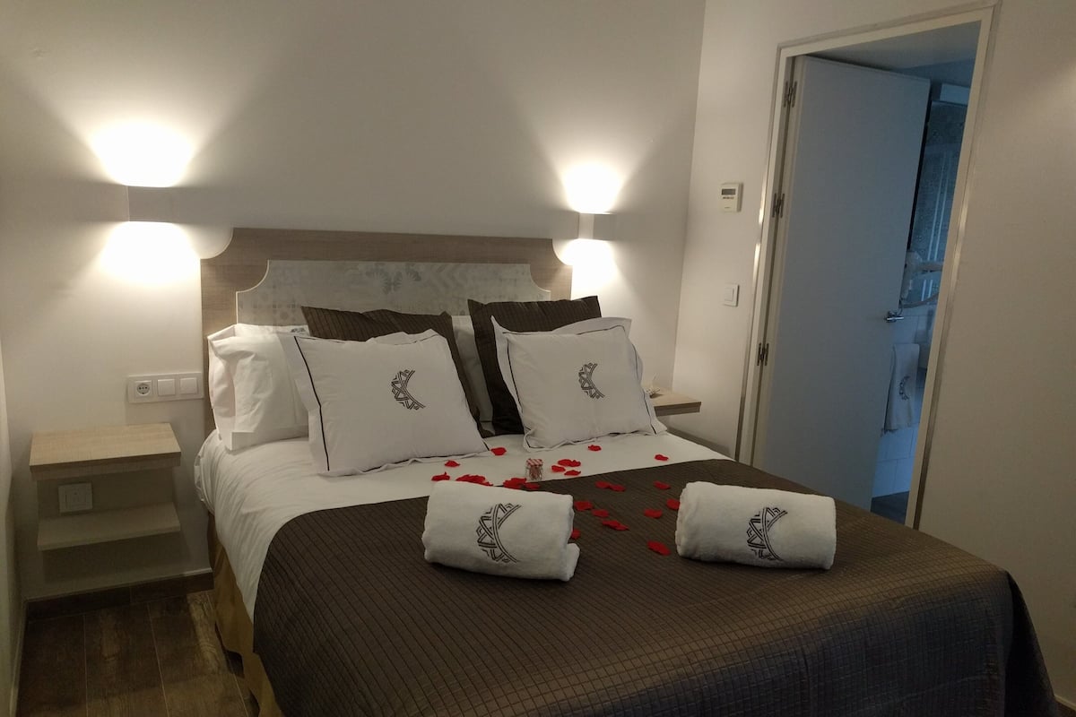 A neatly made bed with a brown bedspread, white pillows with a black design, and two white towels with a black design. There are rose petals on the bed.