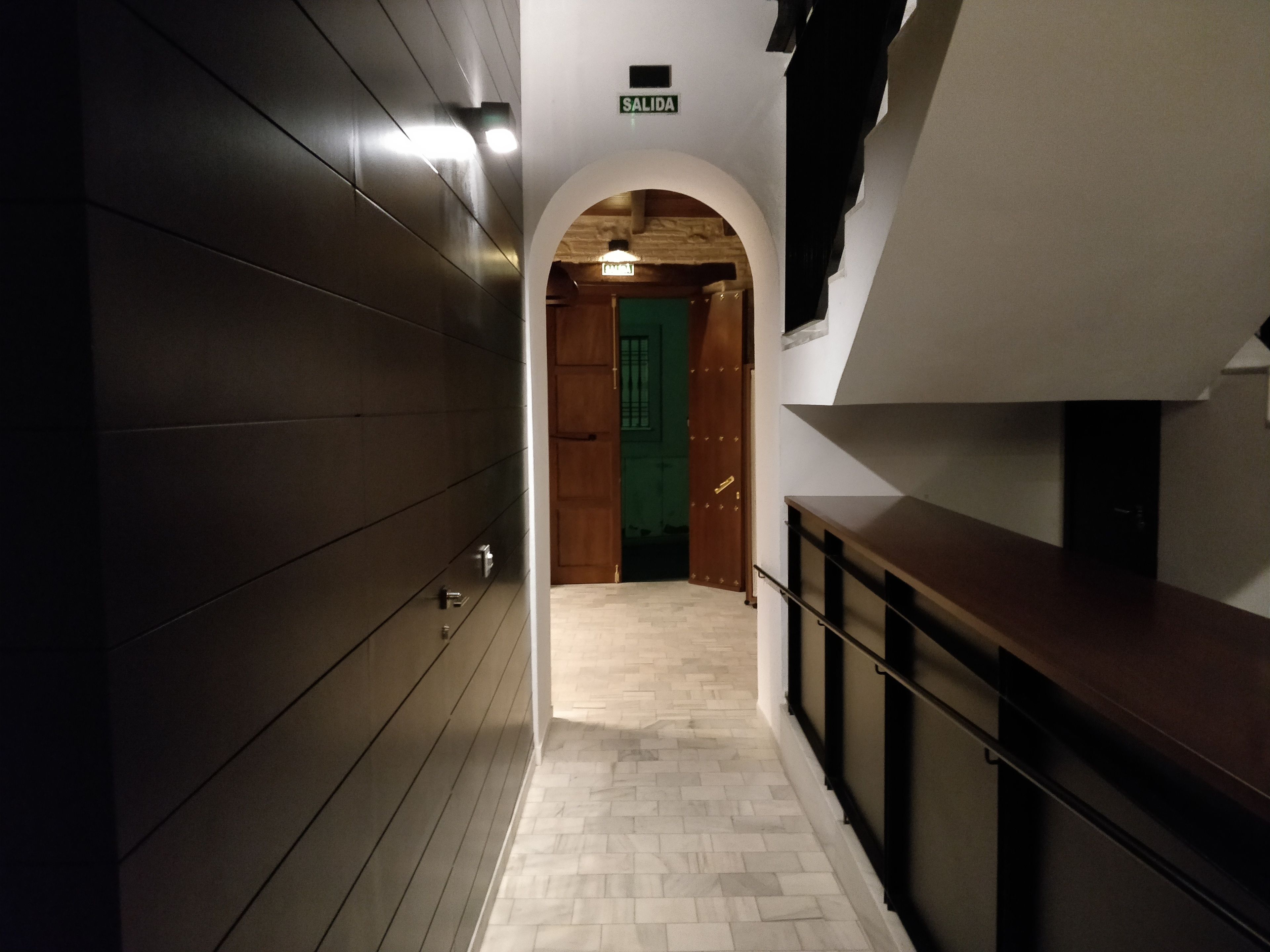 Un couloir se termine par une porte en bois, une enseigne est placée au-dessus, et un escalier se trouve sur la droite.
