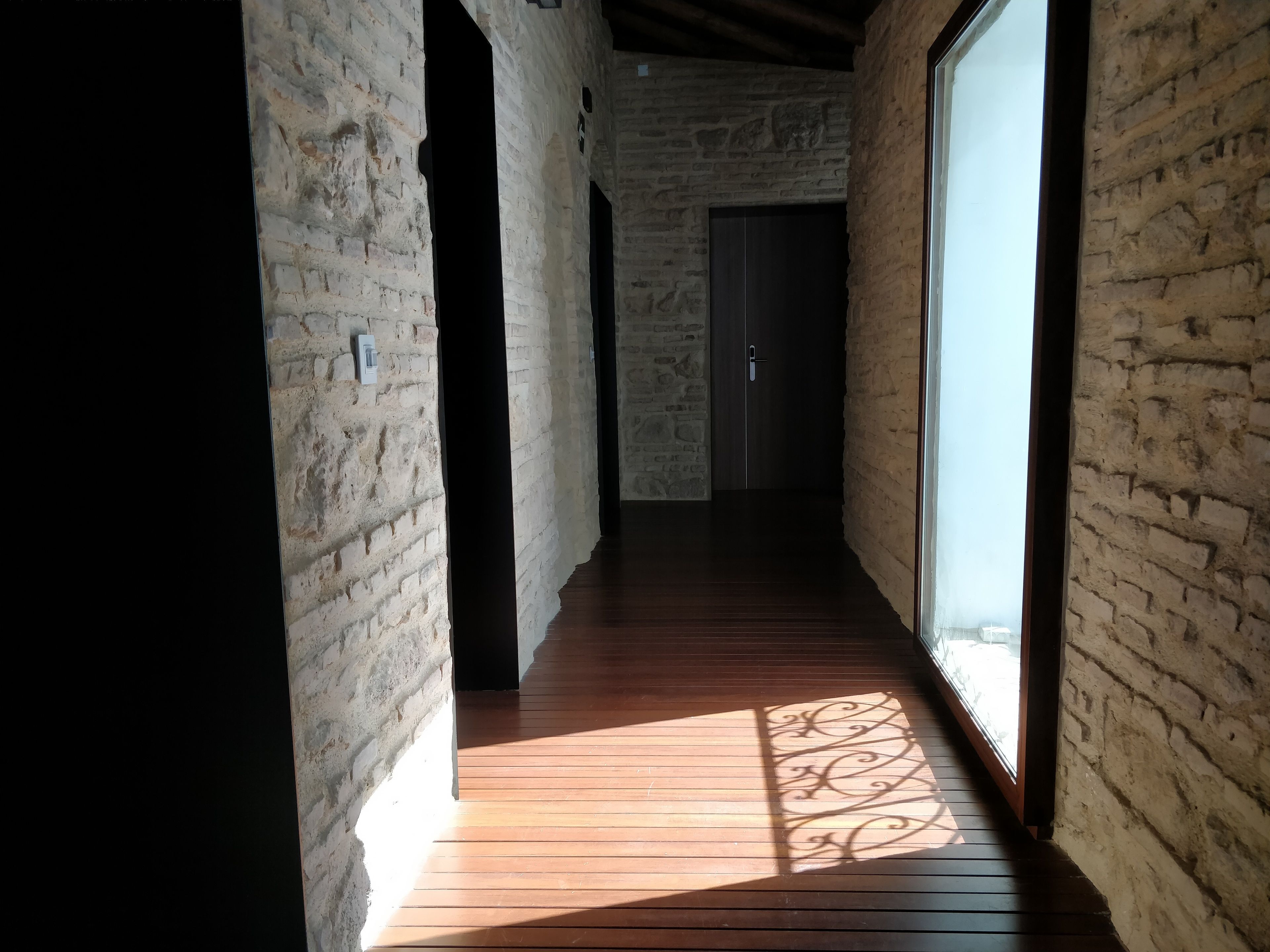 Un couloir étroit aux murs en briques apparentes et au plancher en bois, qui mène à une porte sombre.