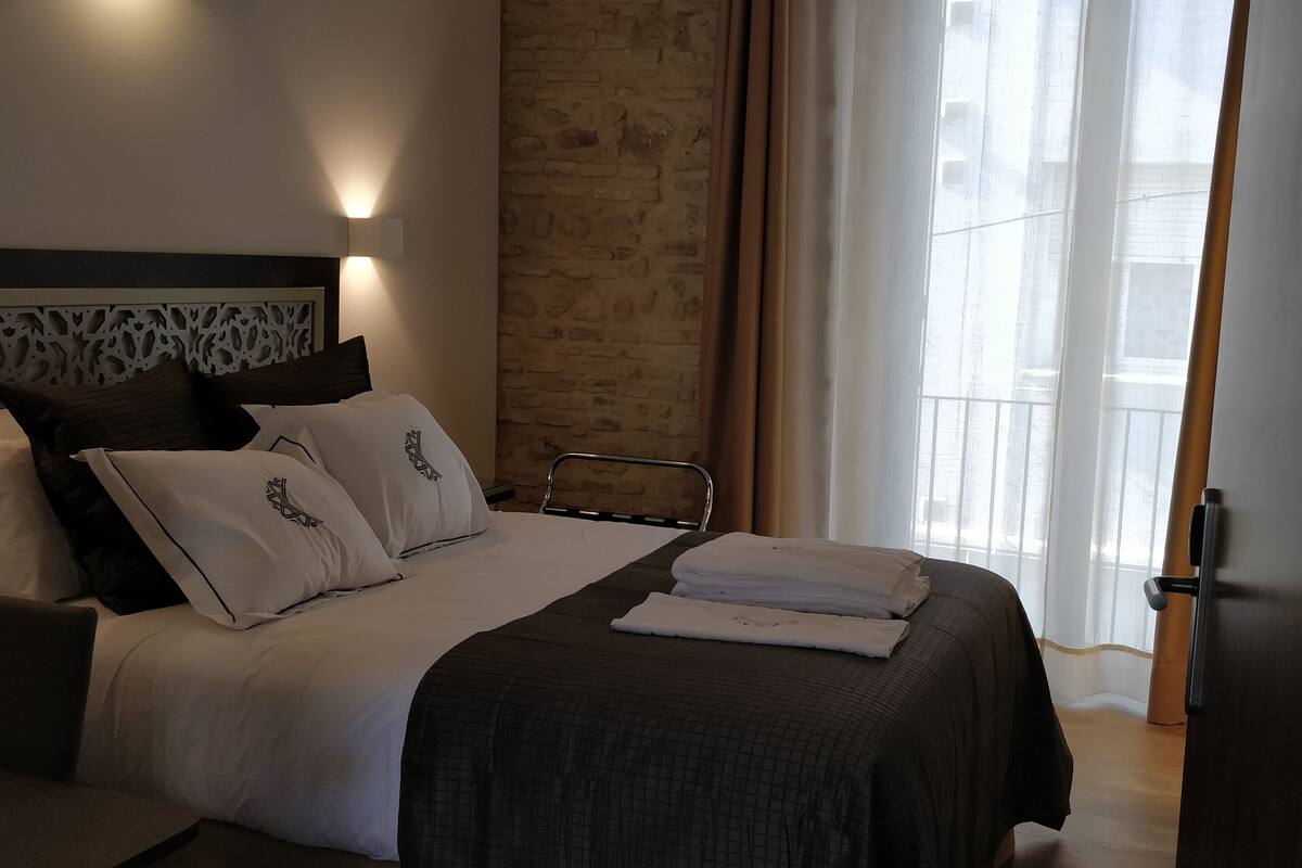 A neatly made bed with white and dark bedding, a headboard with a decorative pattern, and a view of a cityscape through sheer curtains.