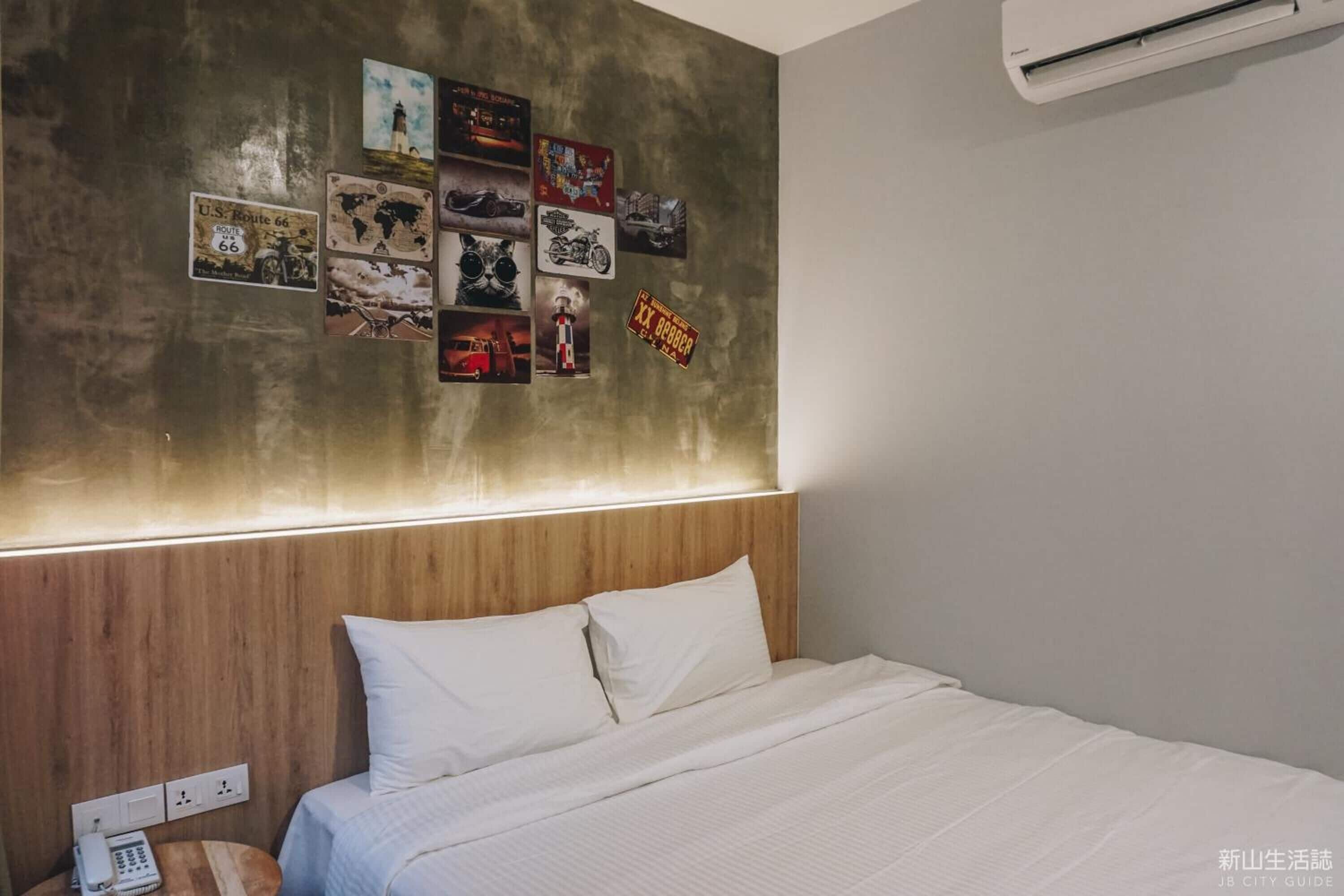 Habitación de hotel con cama, cabecera de madera, pared decorada con carteles y unidad de aire acondicionado.