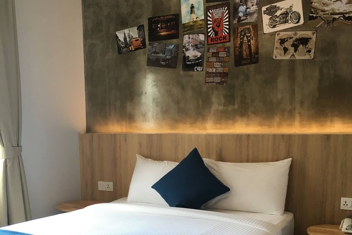 A neatly made bed with white and blue bedding, a blue throw blanket, and two rolled towels. The room has a wooden headboard and a small wooden table beside the bed.