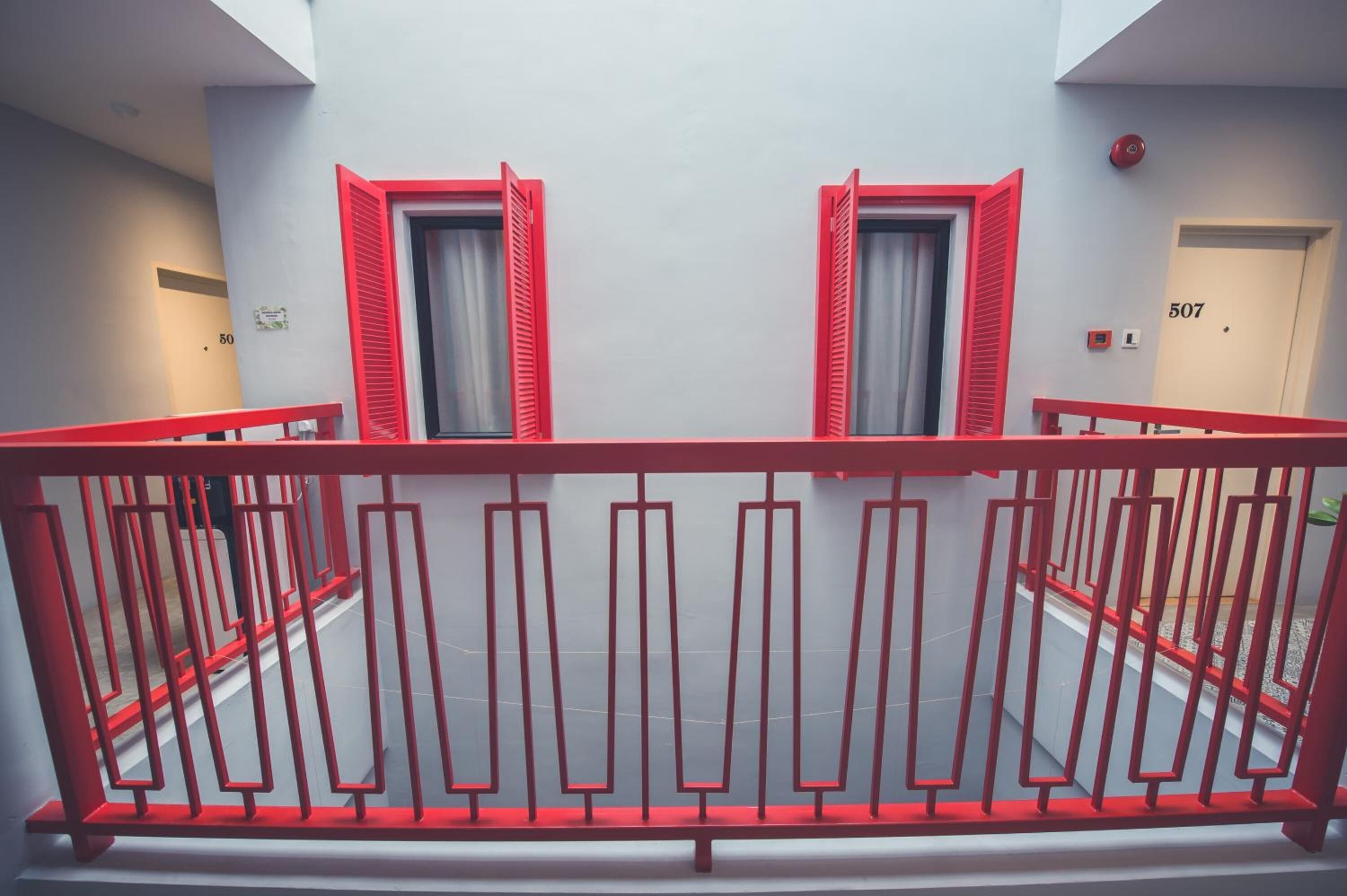 A modern hotel interior with a red railing, two windows with red shutters, and a door marked with the number 507.