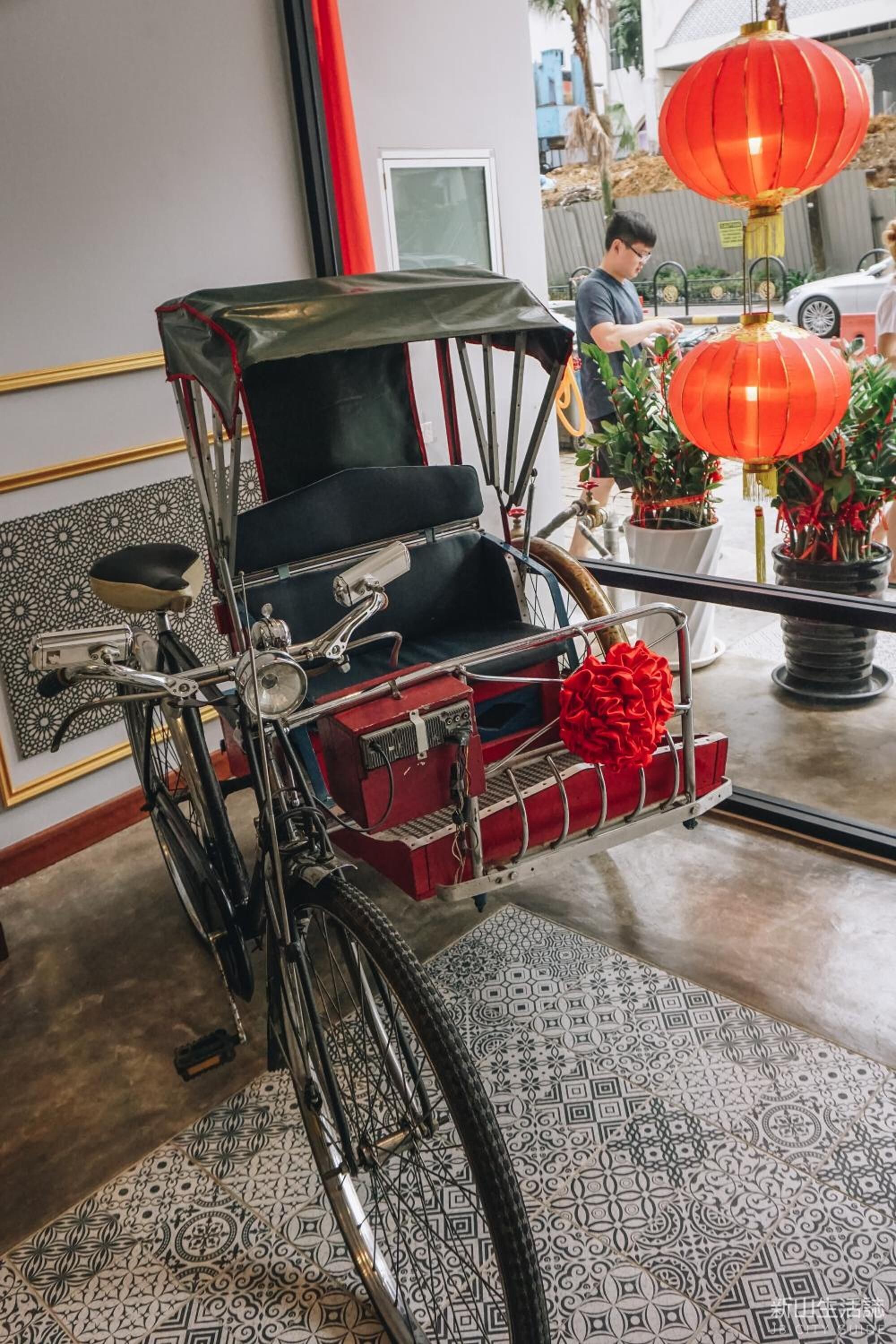 A traditional rickshaw with a black seat and a red box in the front, parked inside a room with patterned floor tiles and a red lantern in the background.