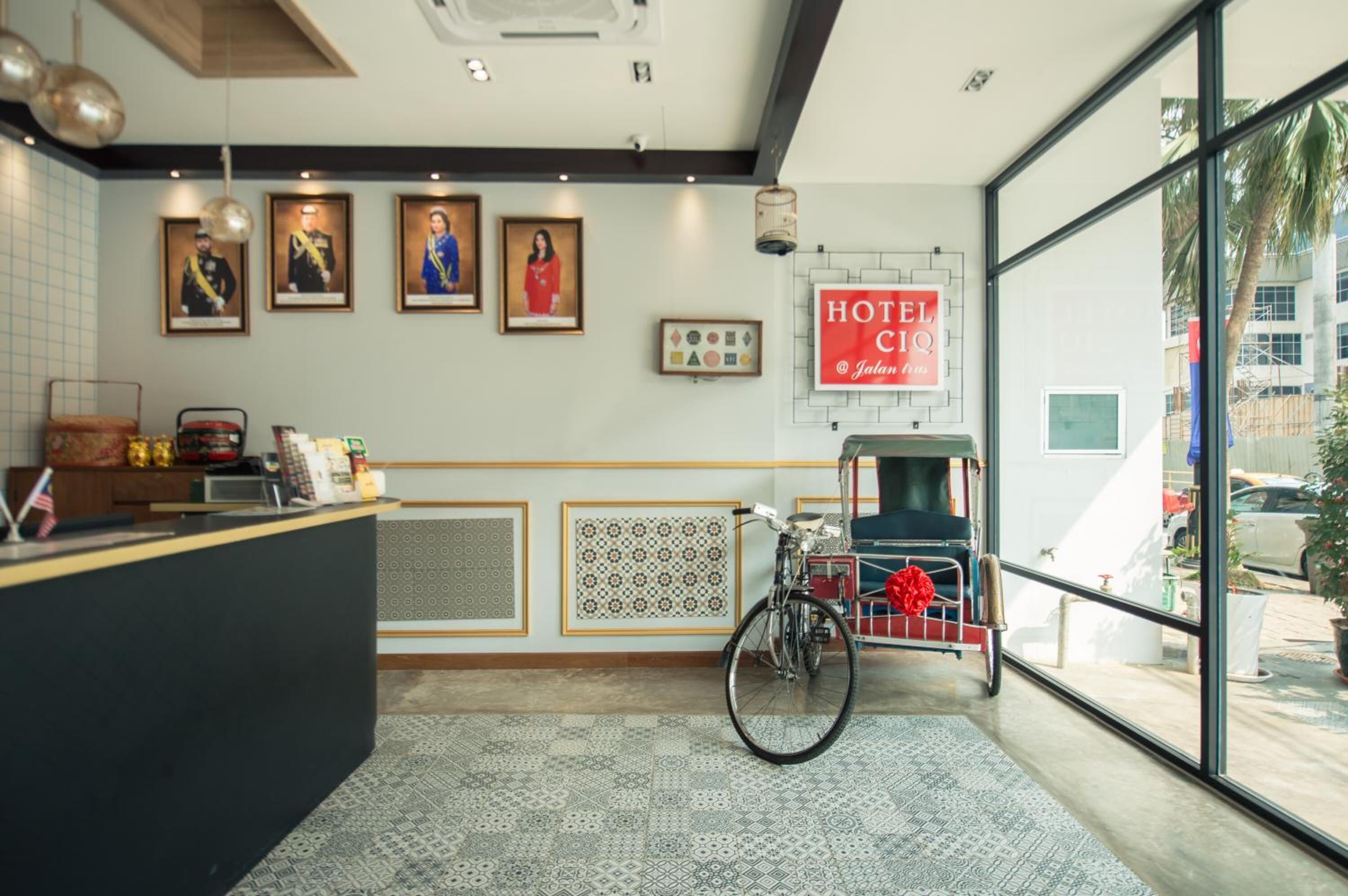 A hotel reception area with a reception desk, framed pictures on the wall, a bicycle, and a large window offering a view of the outside.