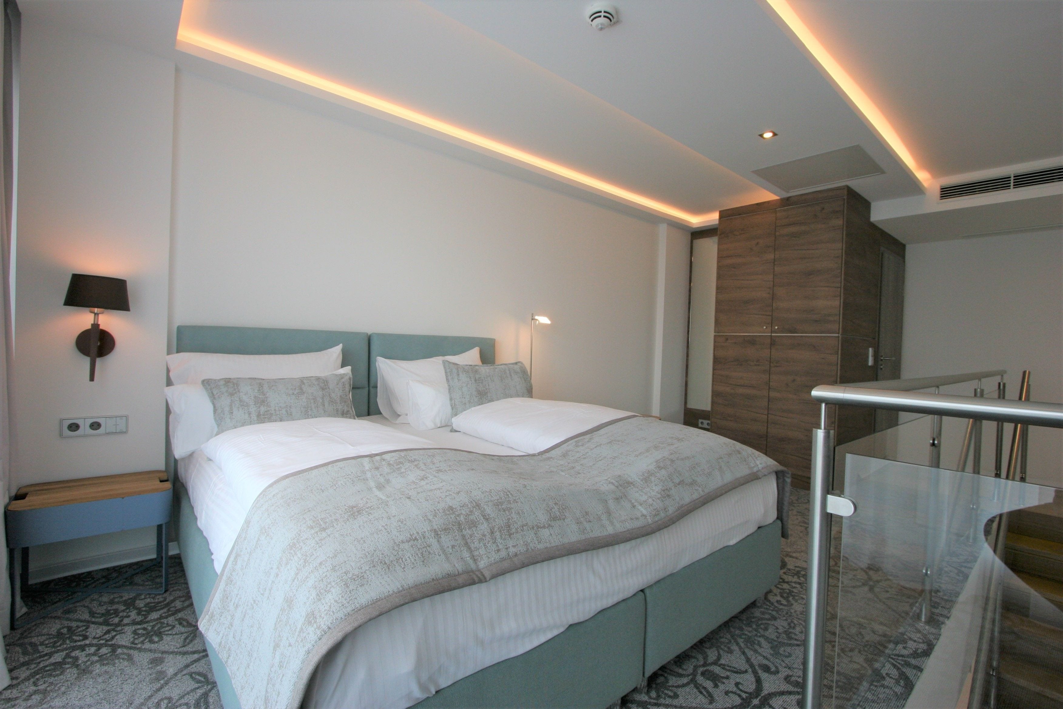 Une chambre d’hôtel moderne dotée d’un grand lit, de tables de chevet, d’une petite table, d’une armoire et d’un escalier en verre.