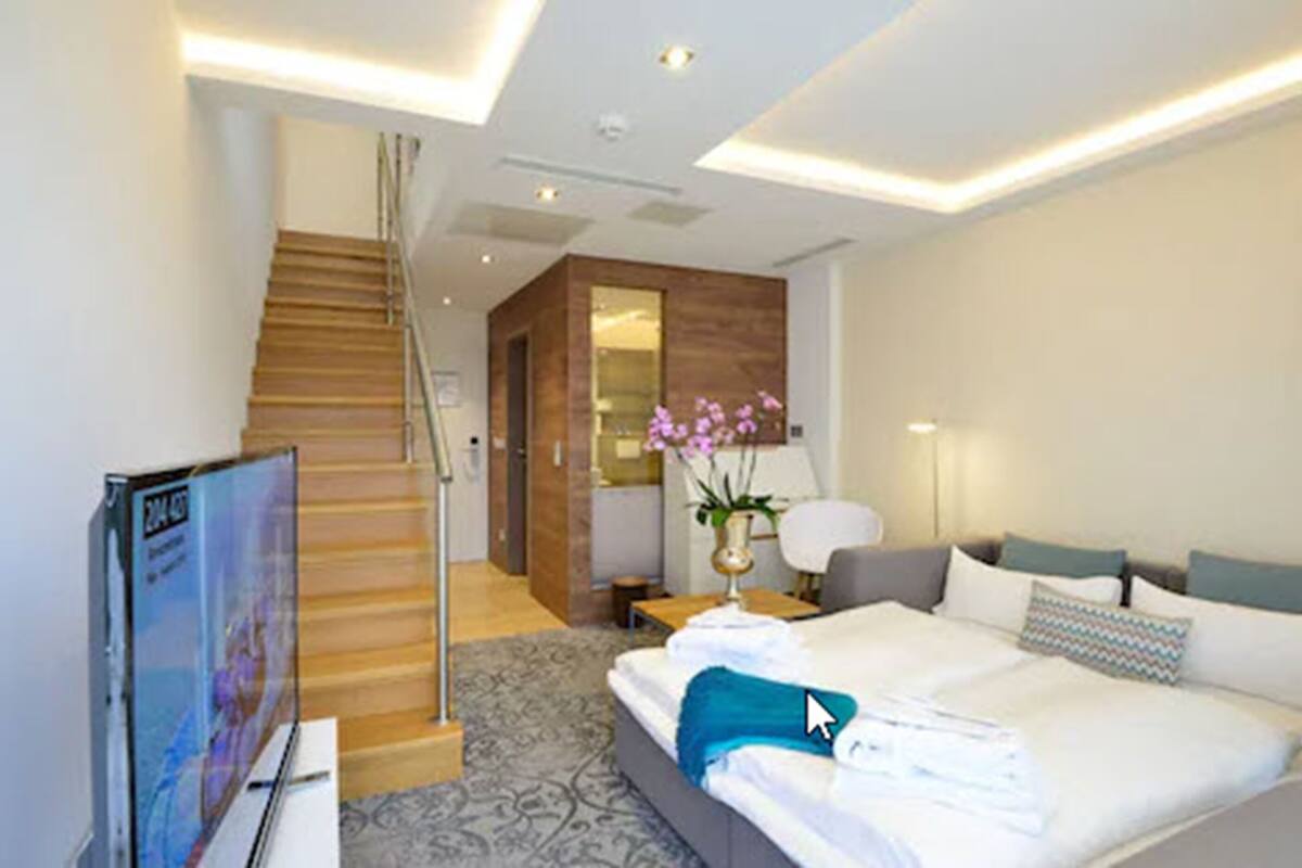 A modern hotel room with a neatly made bed, a flat-screen TV, a wooden staircase, and a small table with a lamp.