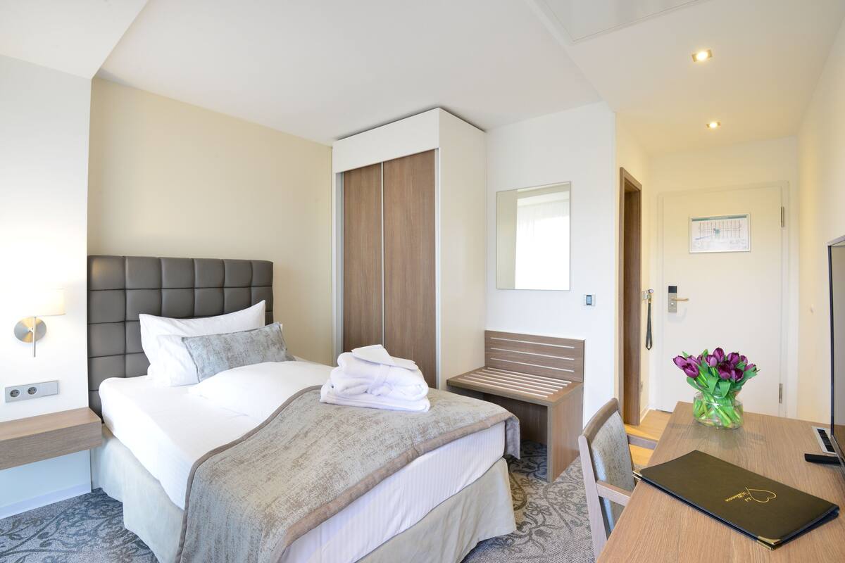 A hotel room with a large bed, a wooden wardrobe, a desk with a laptop, and a vase with flowers.