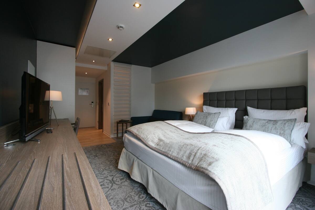 A modern hotel room with a large bed, bedside lamps, a flat-screen TV, and a wooden side table.
