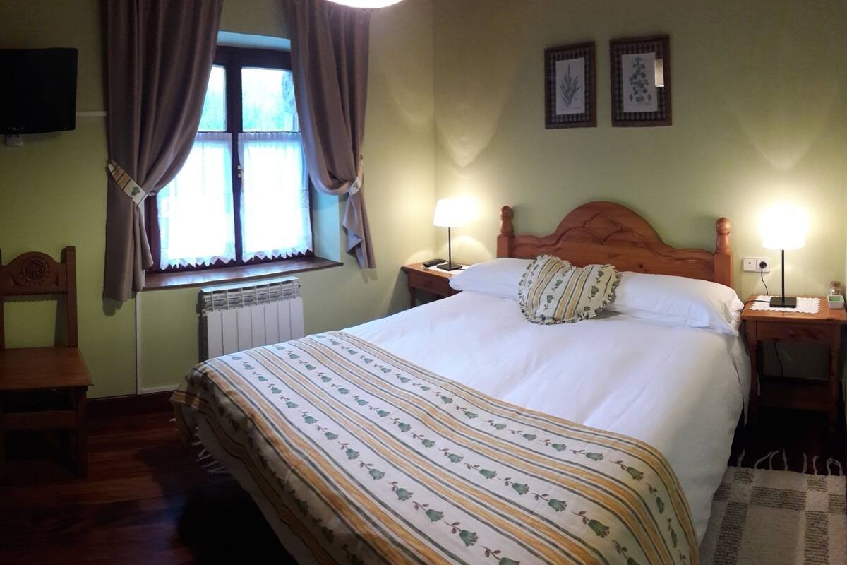 Een netjes opgemaakt bed met een gestreept laken, een houten nachtkastje met een lamp, een stoel en een raam met gordijnen.