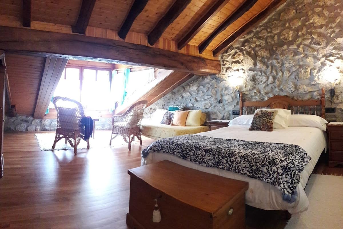 Een gezellige slaapkamer met een stenen muur, houten balkenplafond, een groot bed met beddengoed met een patroon, twee rieten stoelen en een bankje.