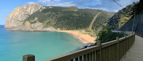 A coastal view with a wooden walkway along the beach, a rocky cliff, and a forested hillside.