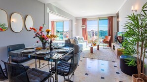 A modern living room with a glass dining table, comfortable seating, a sofa, and a large window offering a city view. - PLAZA TERRACE  AP4065 by RIVIERA HOLIDAY HOMES (Niza)