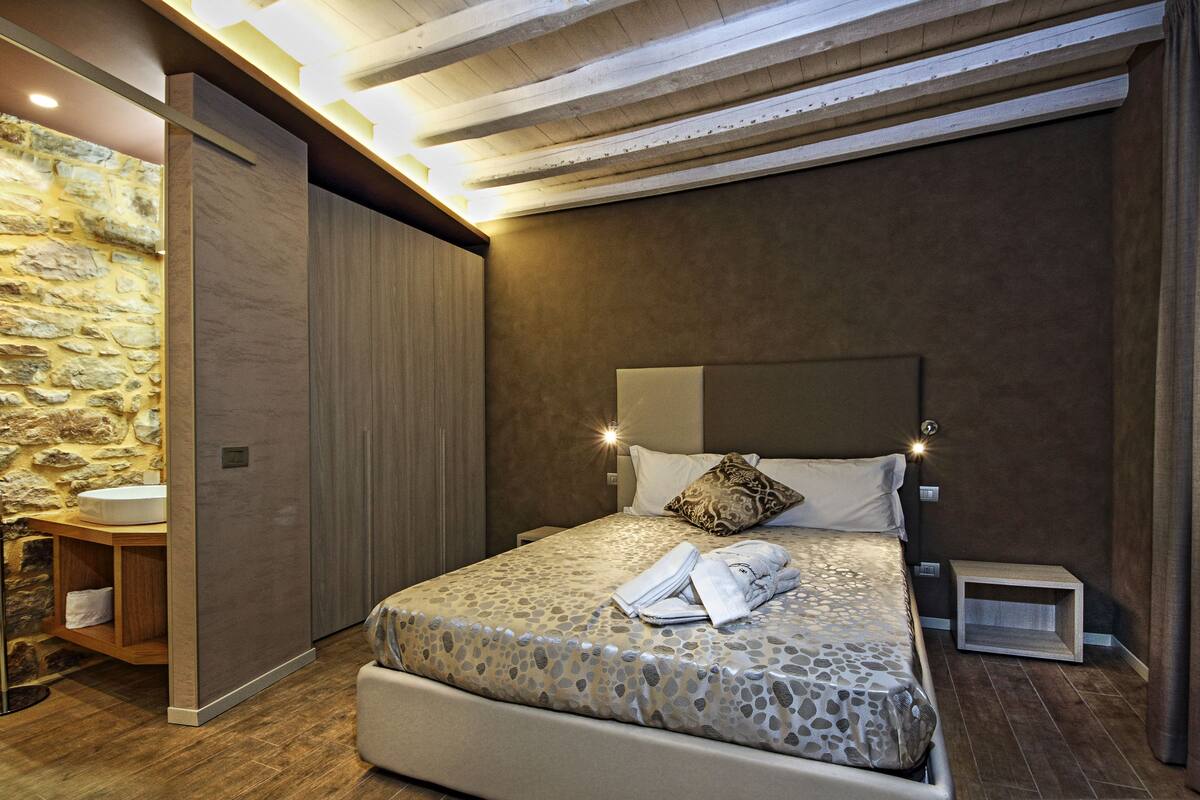 A bedroom with a stone wall, a wooden ceiling, a bed with a patterned bedspread, and a nightstand.