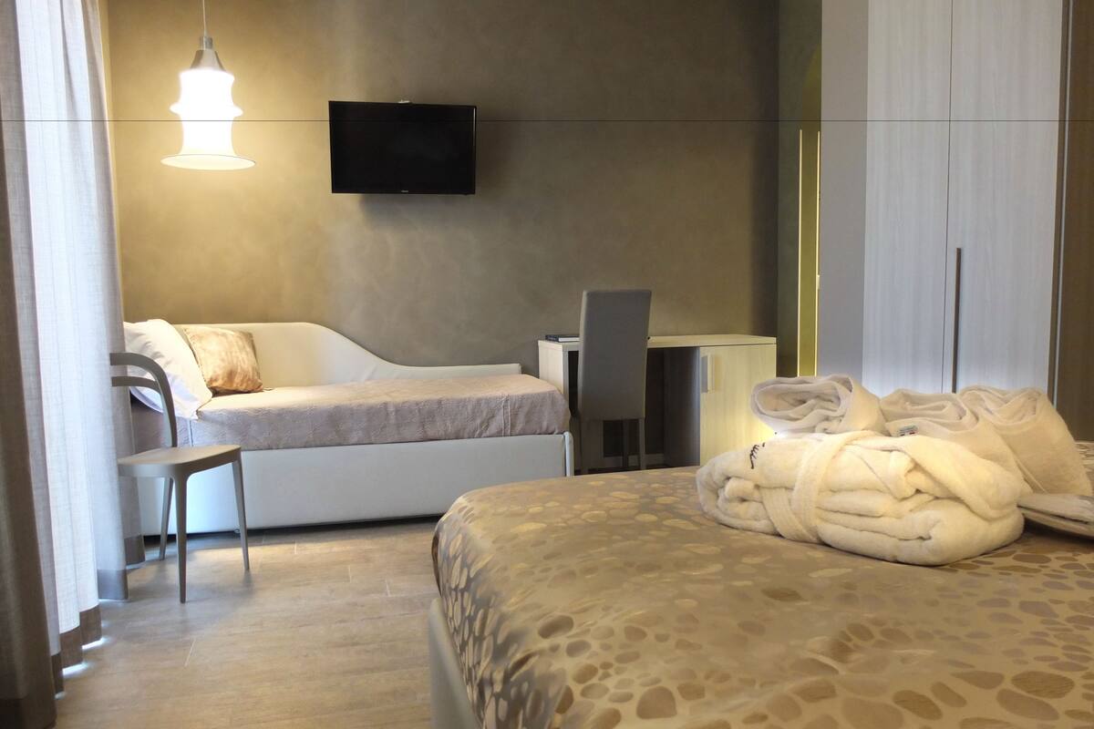 A modern hotel room with a bed, a desk, a chair, a TV mounted on the wall, and a window with curtains.
