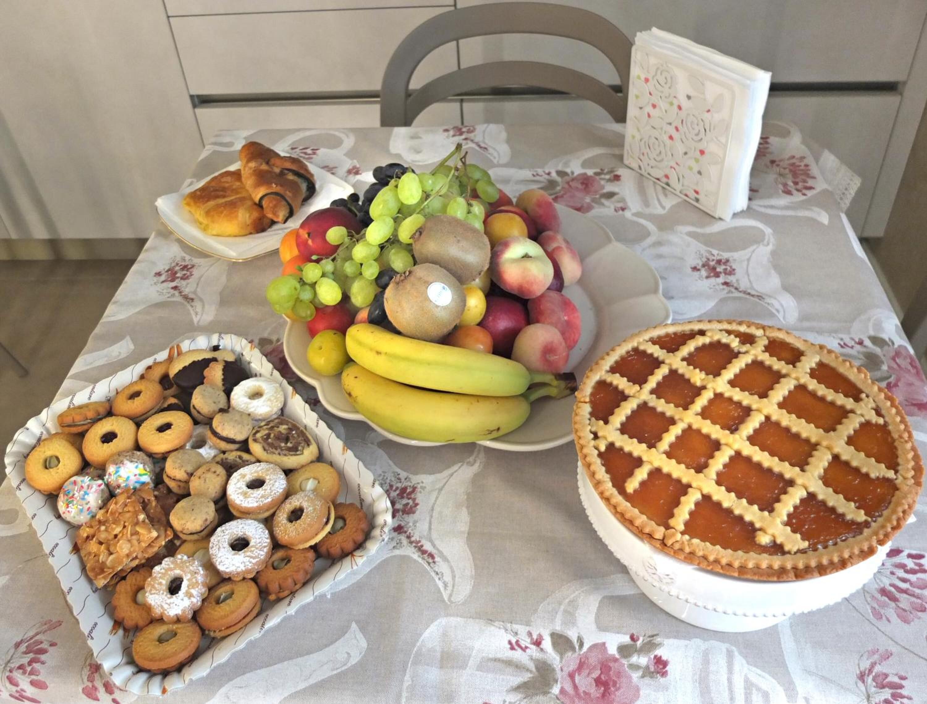 Una mesa con mantel floral, un plato de pasteles, un bol de fruta y un pastel con corteza de rejilla.
