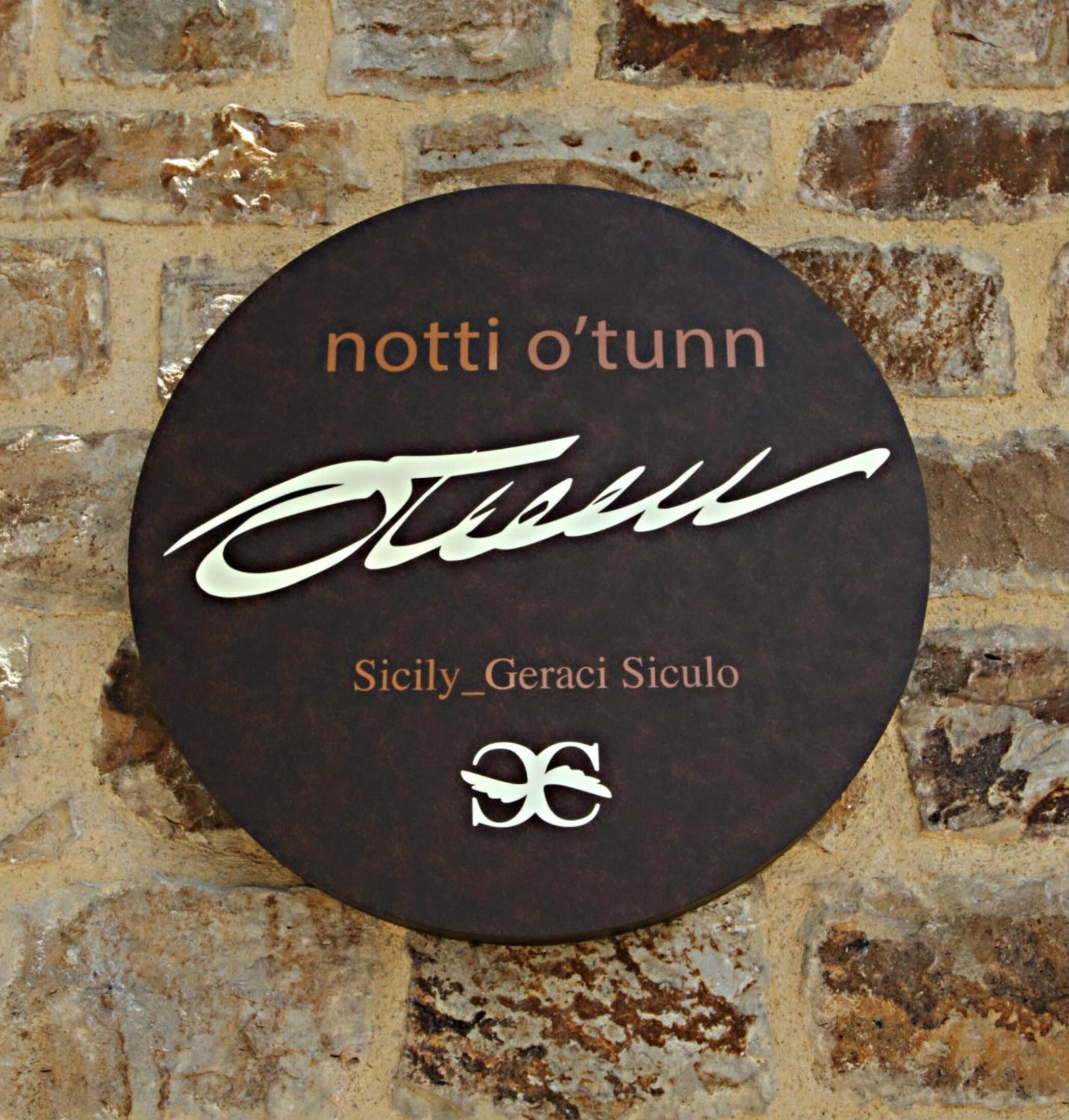A circular sign with the text "notti o'tunn" and "Sicily_Geraci Siculo" against a stone wall background.