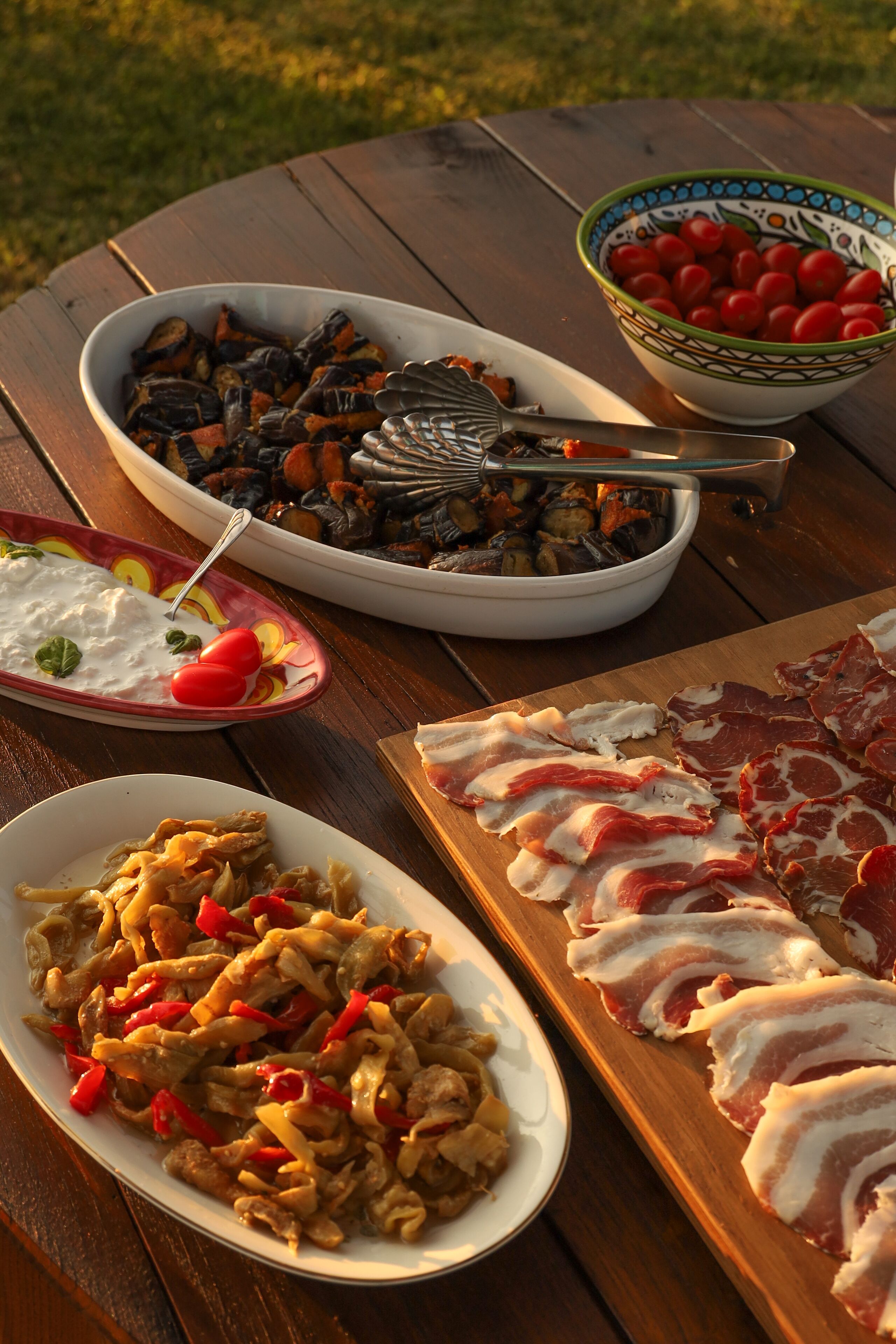Un tavolo di legno con diversi piatti, tra cui verdure grigliate, carne a fette e una ciotola di pomodorini.