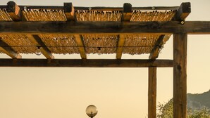 A wooden pergola with a thatched roof, wooden benches, and a large barrel in a garden setting with trees and hills in the background. - L' Aia Resort (Trentinara)