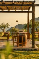 Una pérgola de madera con techo de paja, bancos de madera y un barril grande en un jardín con árboles y colinas al fondo.