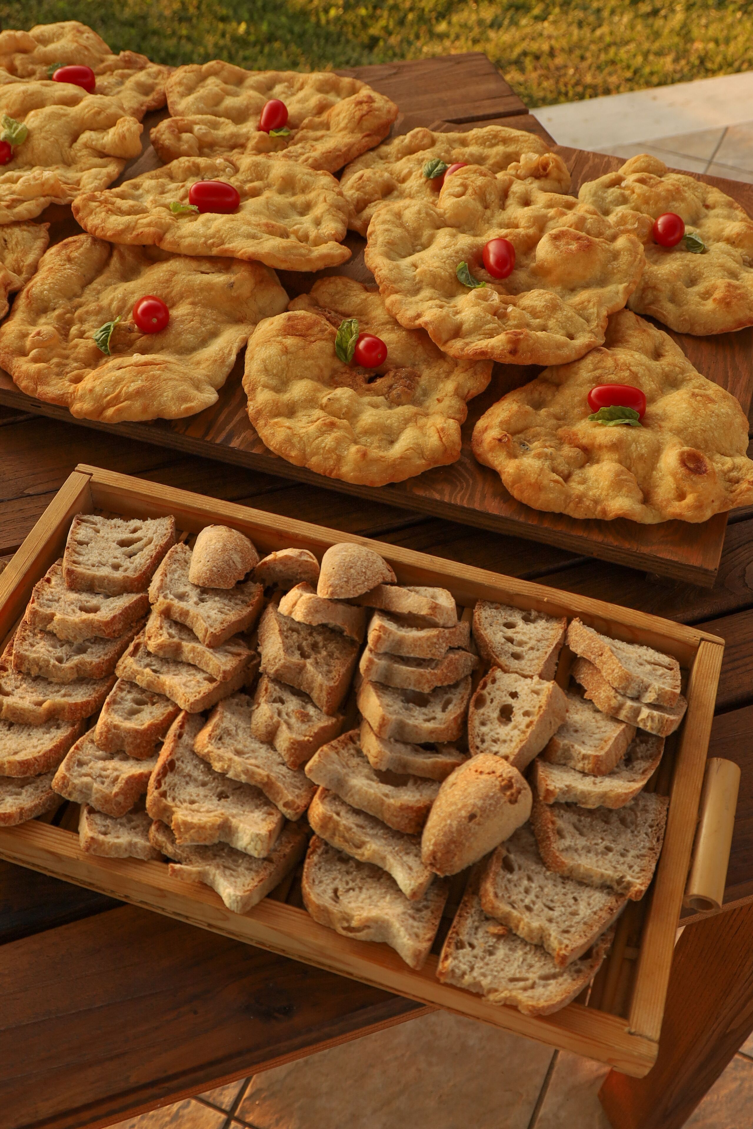 A wooden table with a tray of sliced bread and a wooden board with round, golden-brown flatbreads garnished with cherry tomatoes.