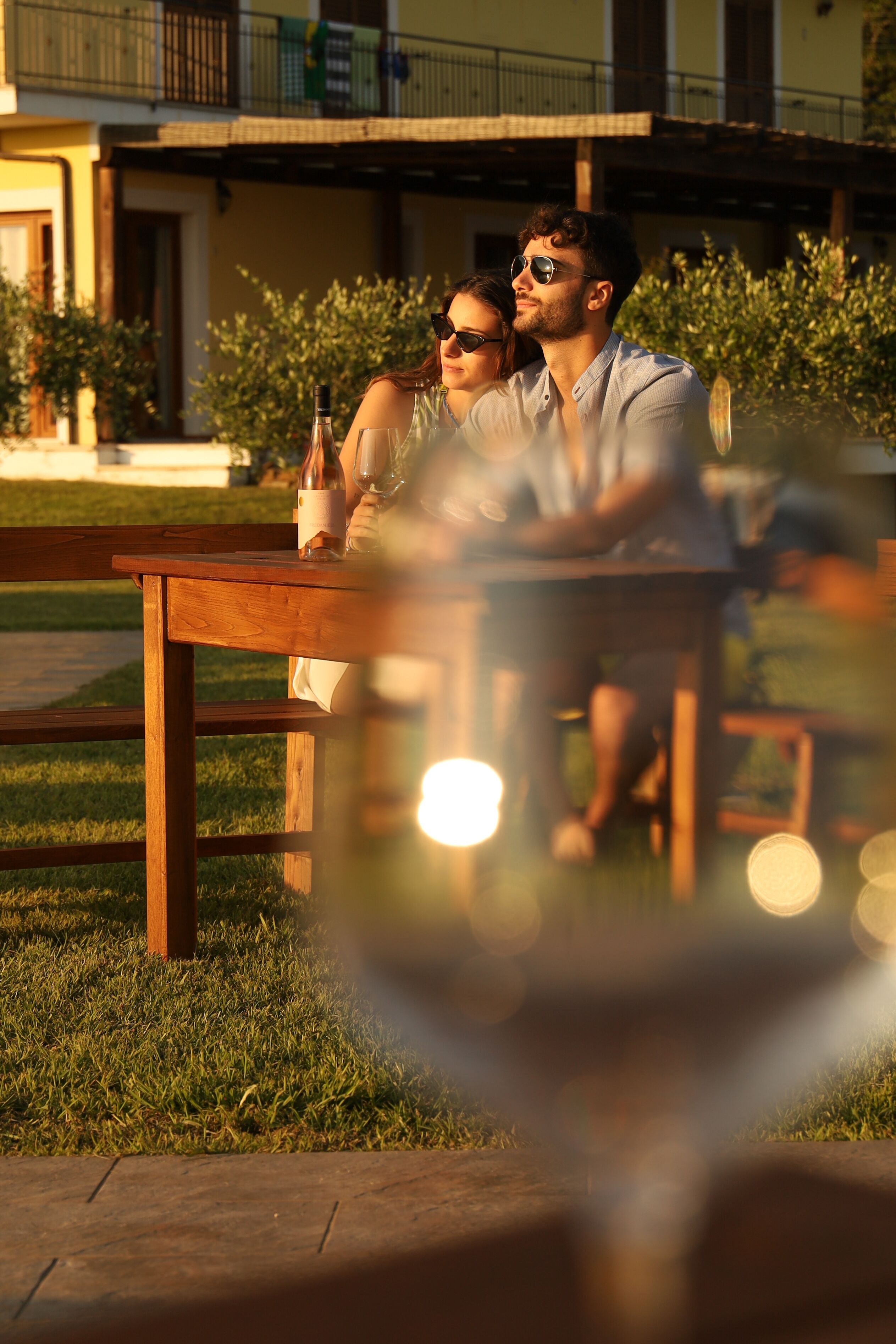 A couple sitting at an outdoor wooden table with a bottle and glass, surrounded by greenery and a building in the background.