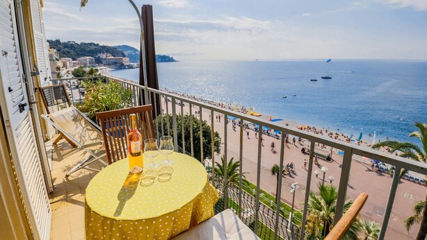 Un balcón con un mantel amarillo de lunares, una botella de vino y copas. Se ve una playa con gente y barcos.