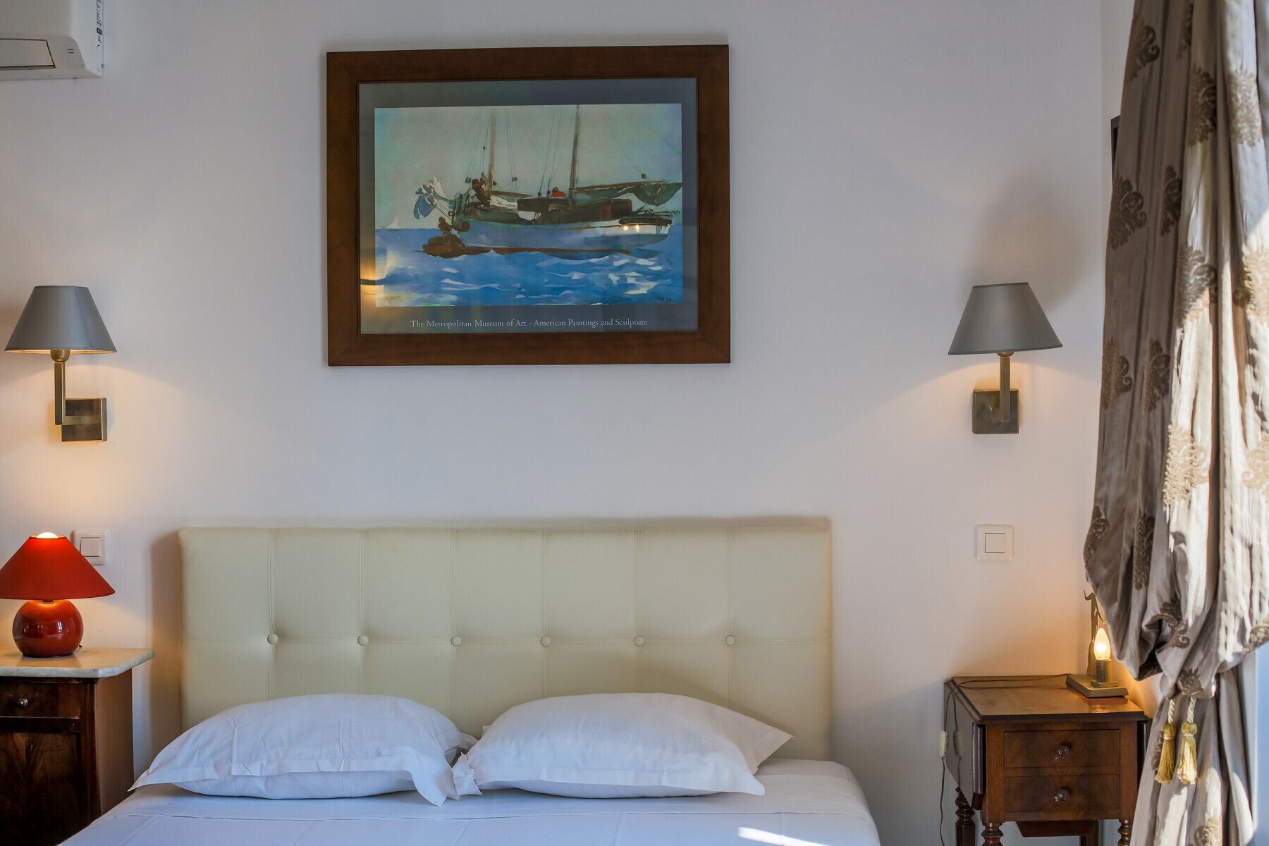 Ein Hotelzimmer mit Bett, zwei Nachttischlampen, einem Segelbootgemälde und einem Fenster mit Vorhängen.