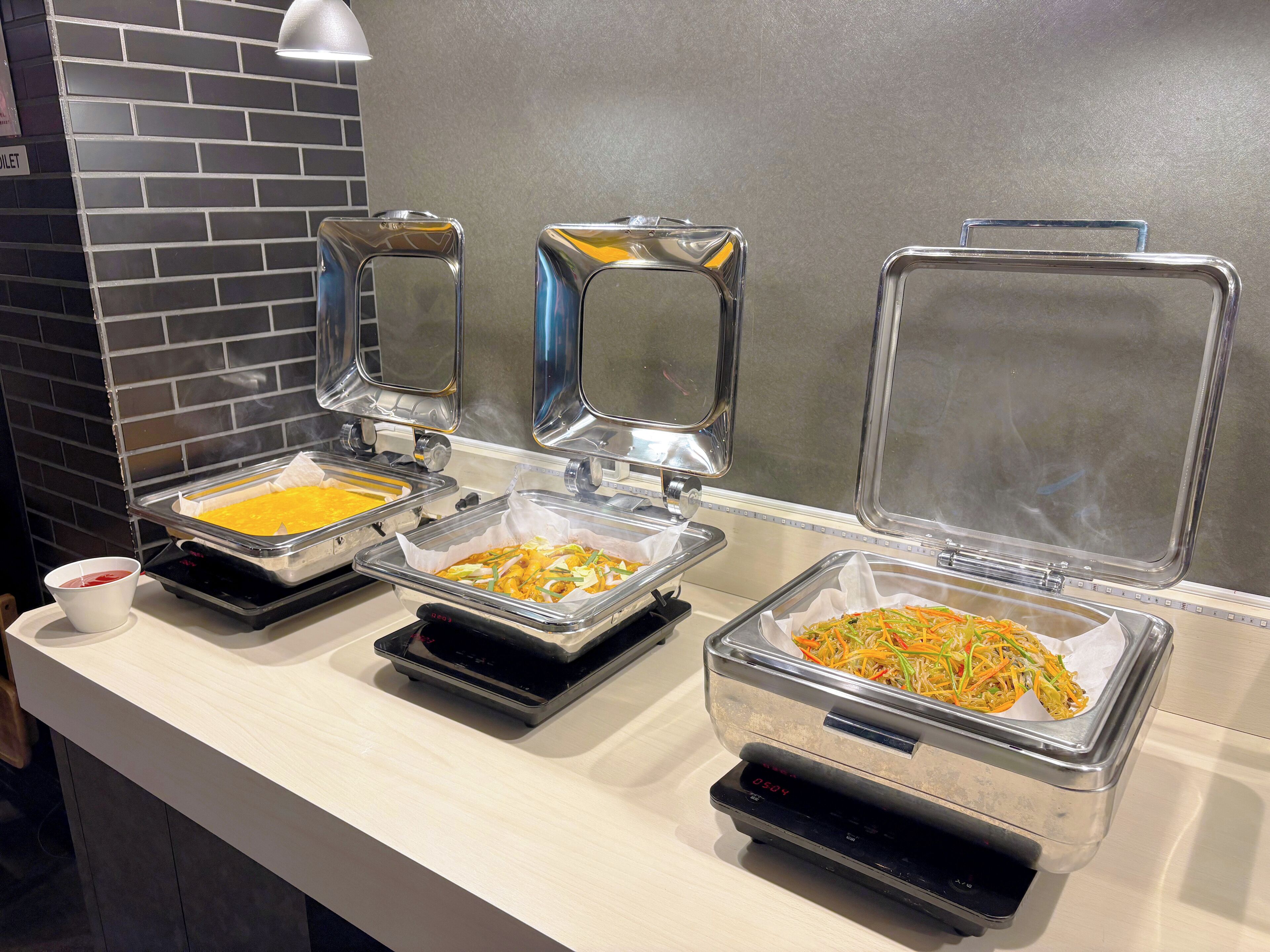 A buffet setup with various food items in transparent containers, a digital scale, and a sign indicating a toilet nearby.